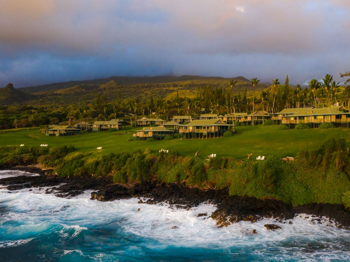 A scenic coastline with lush greenery, buildings set back from the shore, cliffs, and ocean waves crashing against the rocks under a cloudy sky.