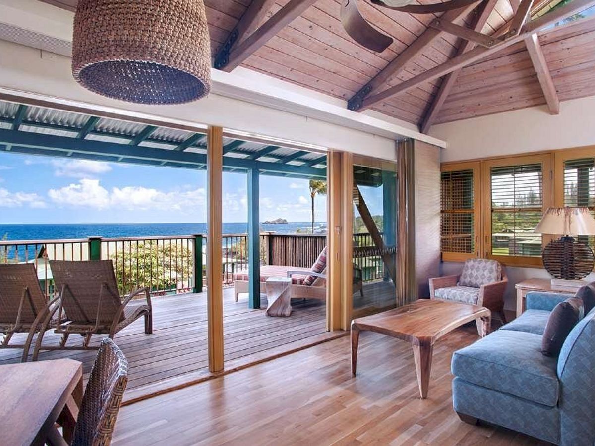 A cozy living room with wooden floors and ceiling, opening to a balcony with sea views, featuring furniture, a hanging lamp, and a ceiling fan.