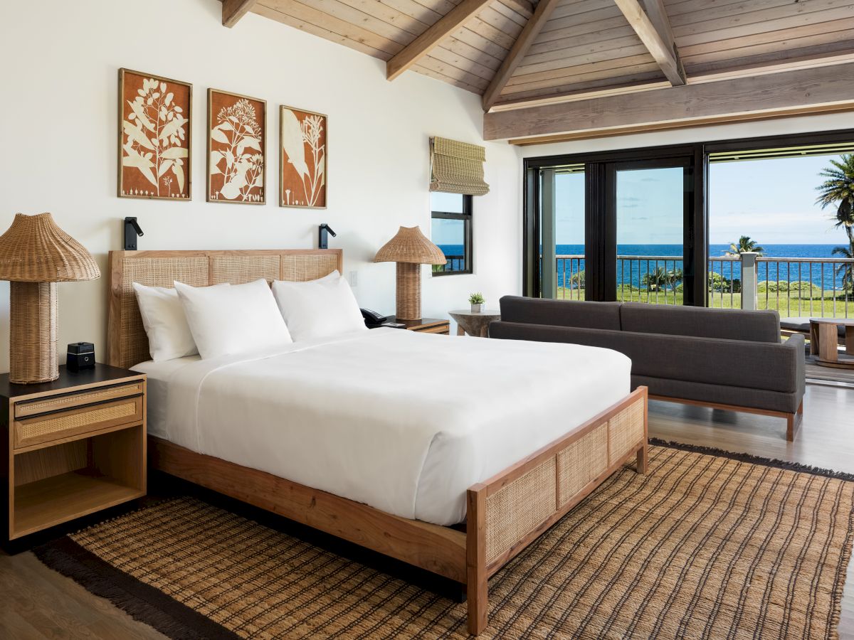 A cozy bedroom with a large bed, wooden furnishings, and a view of the ocean through large windows. décor includes art and a woven rug.