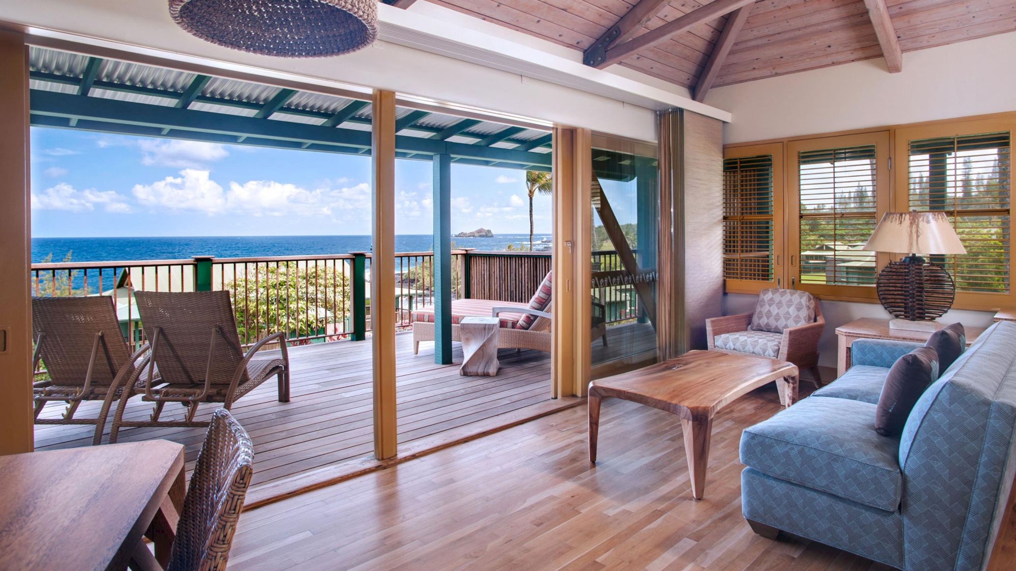 This image shows a cozy living room with wooden flooring, a sofa, chairs, and a coffee table, leading to a balcony with an ocean view.