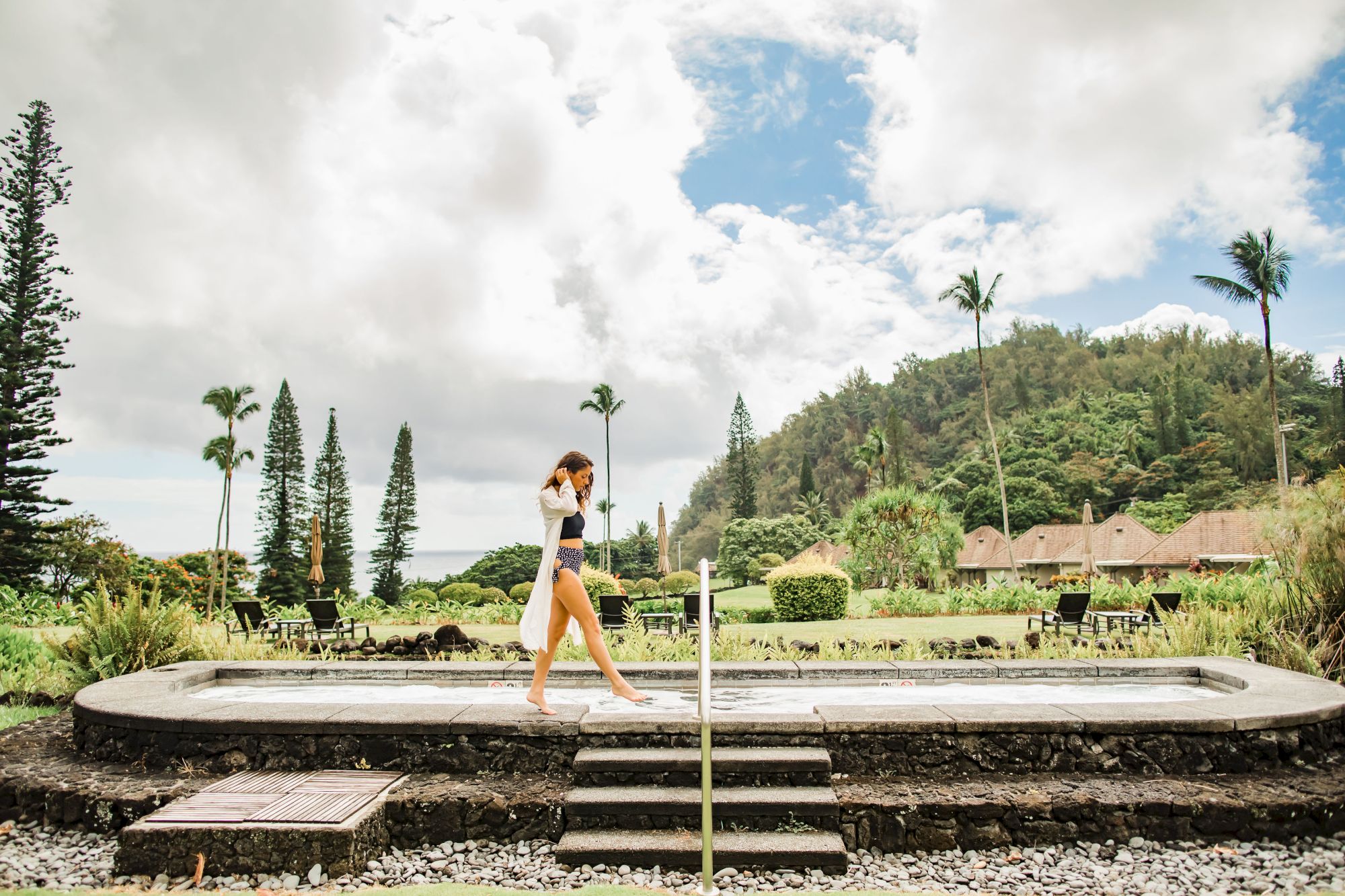 A person walks by an outdoor pool in a lush, scenic area with mountains, palm trees, and greenery under a cloudy sky.