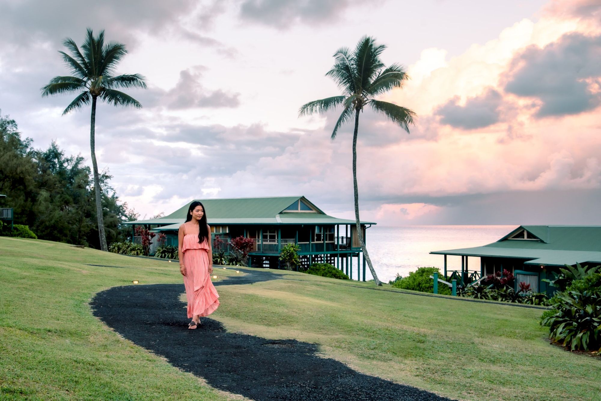 A woman in a pink dress walks on a path through a grassy area with palm trees and cabins, with a scenic ocean view in the background.