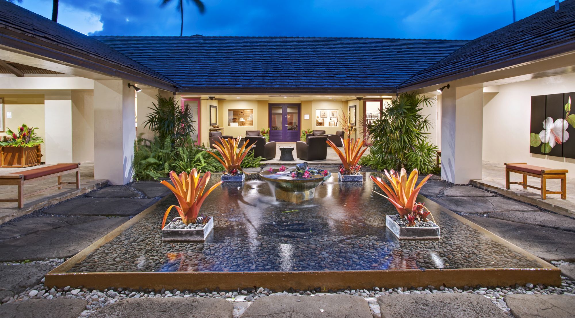 A courtyard with a central water feature and lush plants, surrounded by benches and leading to an inviting interior space through an open doorway.