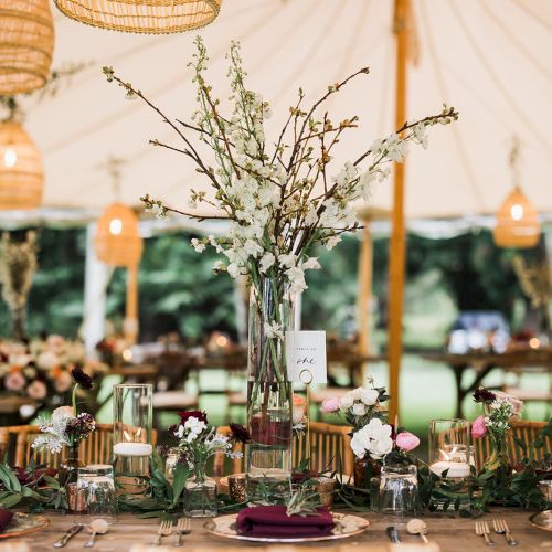 The image shows a beautifully set wedding or event table under a tent with floral centerpieces and hanging lanterns, creating an elegant atmosphere.