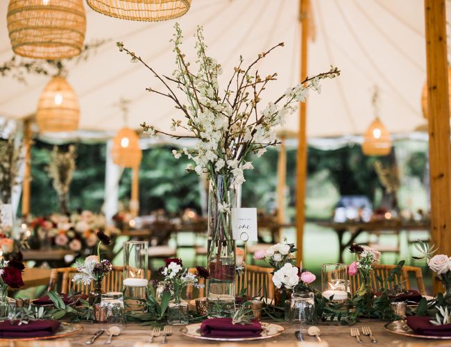 The image shows a beautifully set wedding or event table under a tent with floral centerpieces and hanging lanterns, creating an elegant atmosphere.