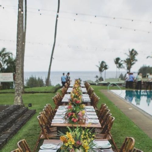 A long, elegantly set dining table with floral arrangements by a pool in an outdoor tropical setting, palm trees, and ocean in the background.