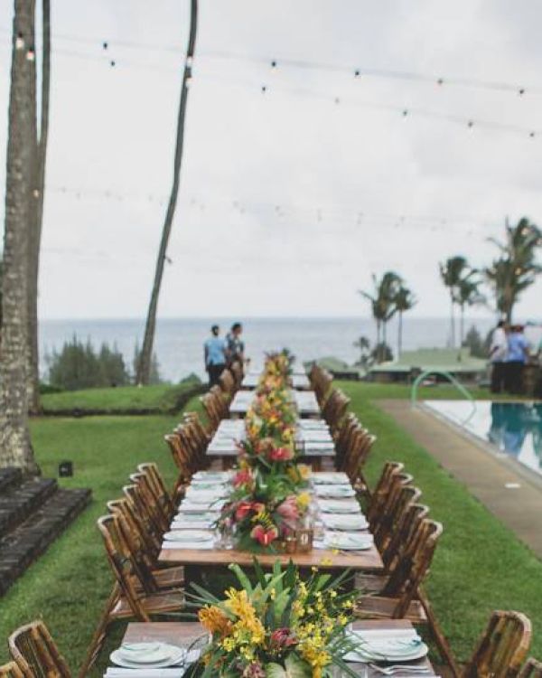 A long, elegantly set dining table with floral arrangements by a pool in an outdoor tropical setting, palm trees, and ocean in the background.