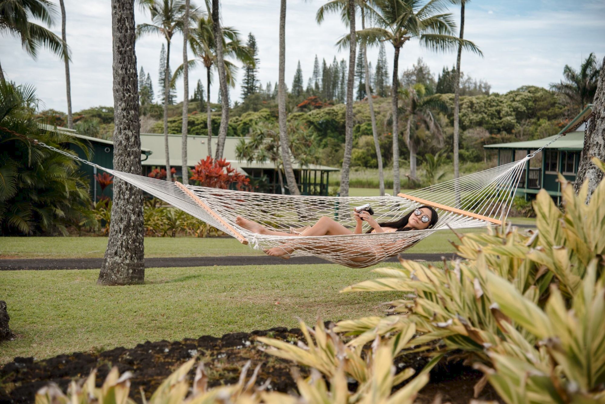 A person is relaxing in a hammock strung between palm trees, surrounded by tropical plants and cottages in the background.