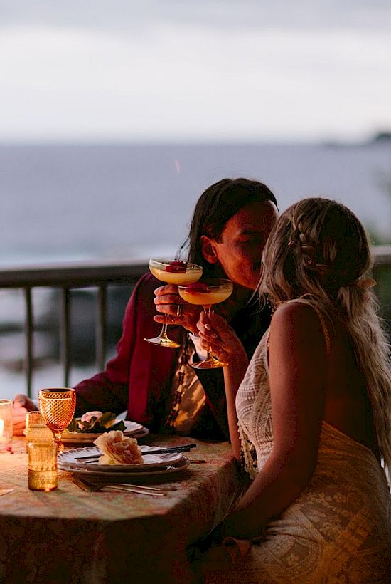 A couple is sitting at a candlelit table near a seaside, holding drinks and engaging in a romantic moment at sunset.