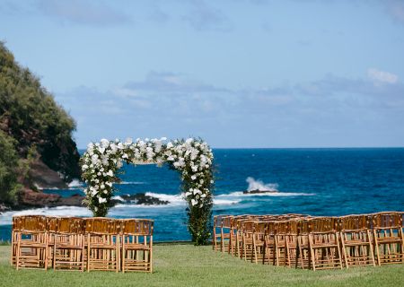 An outdoor wedding setup with a floral arch and rows of wooden chairs facing the ocean, set against a scenic coastal backdrop ending the sentence.