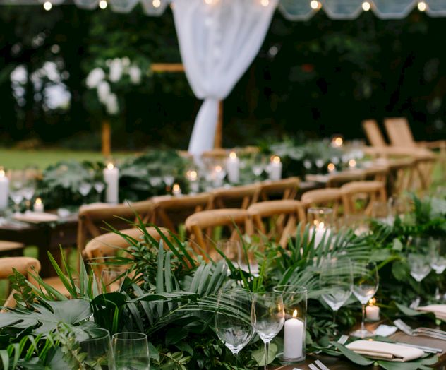 An outdoor event setup with a long table adorned with greenery, wine glasses, candles, and wooden chairs under a transparent tent with string lights.