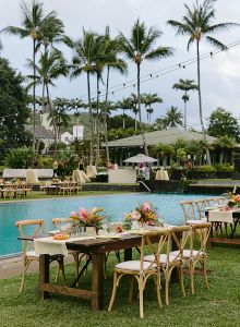 An outdoor setting with a pool, tables, chairs, palm trees, and string lights, suggesting a tropical event or gathering.