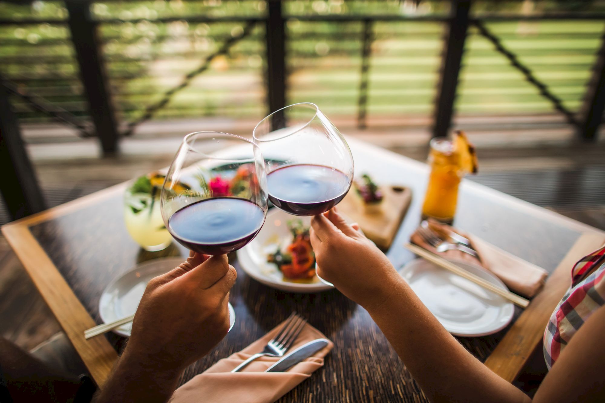 Two people clinking glasses of red wine at a table set for a meal with food and drinks, enjoying a scenic outdoor dining experience.