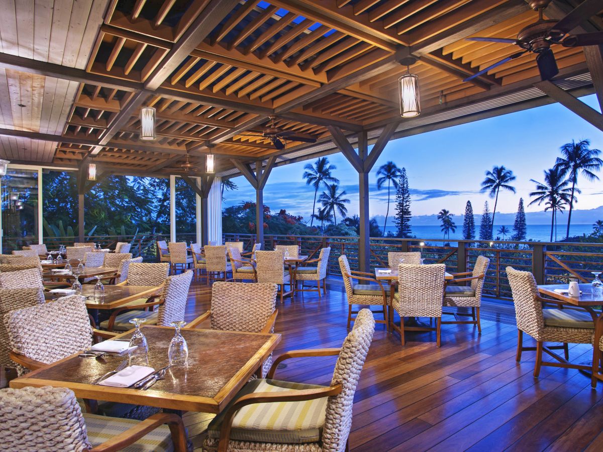 An outdoor dining area with wooden flooring and wicker chairs, overlooking a scenic view of palm trees and the ocean during sunset or early evening.