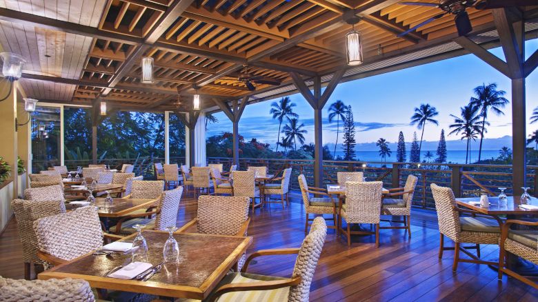 An outdoor dining area with wooden flooring and wicker chairs, overlooking a scenic view of palm trees and the ocean during sunset or early evening.