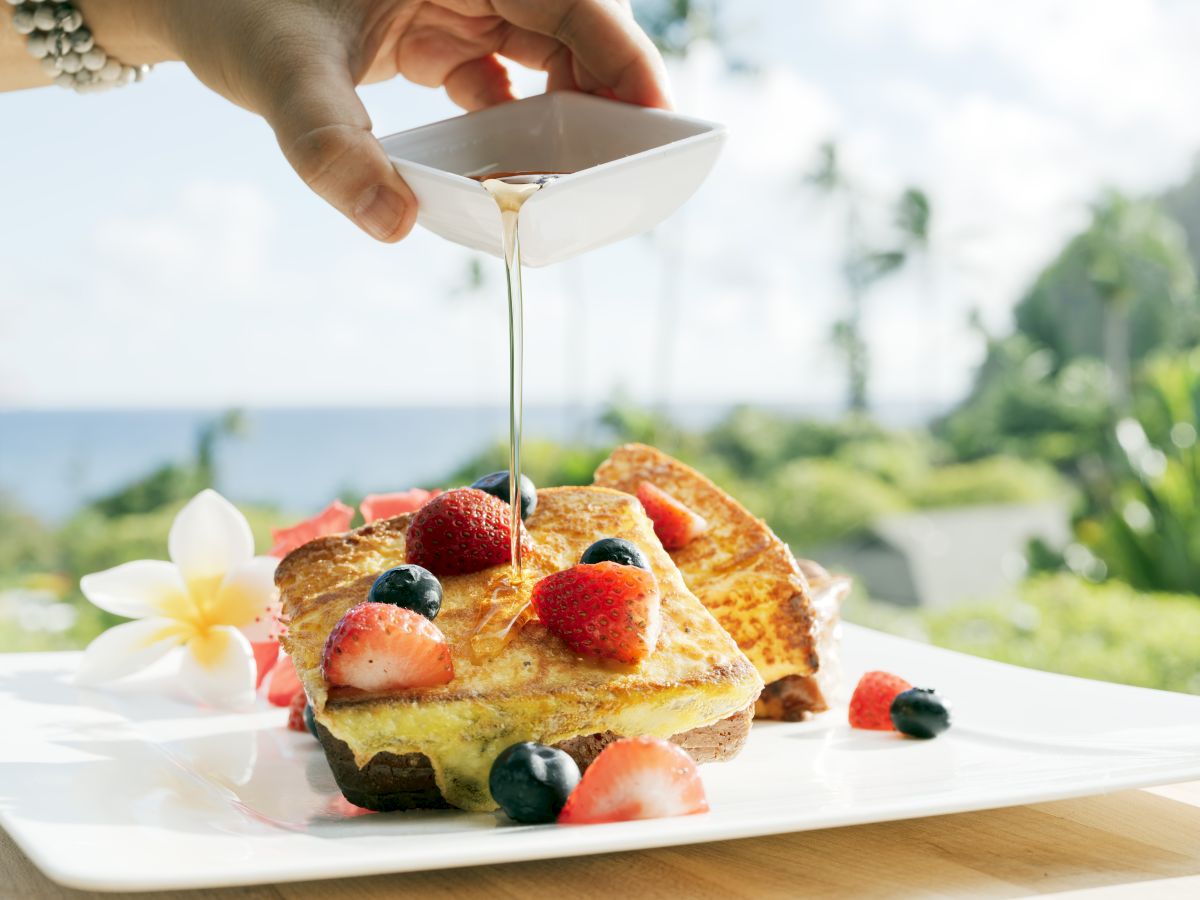 The image shows syrup being poured onto a stack of French toast topped with strawberries and blueberries on a plate.