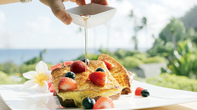 The image shows syrup being poured onto a stack of French toast topped with strawberries and blueberries on a plate.