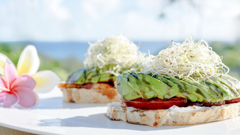 The image shows two slices of bread topped with avocado, tomato, and sprouts on a white plate, near a plumeria flower in a bright setting.