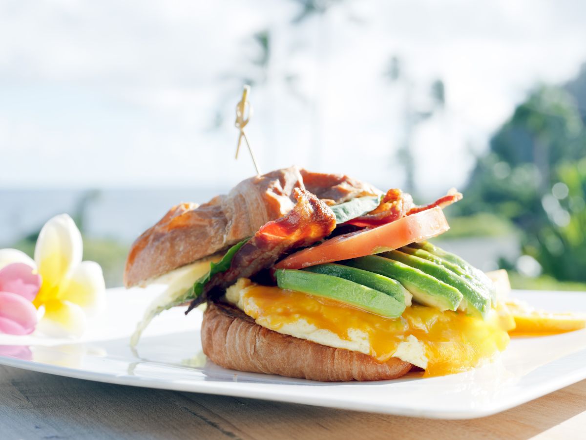The image shows a sandwich with bacon, avocado, cheese, and tomato, placed on a white plate with flowers, set outside in a sunny location.