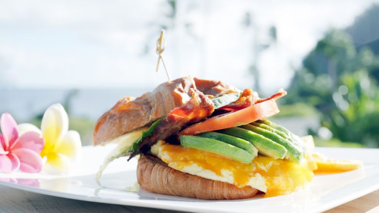 The image shows a sandwich with bacon, avocado, cheese, and tomato, placed on a white plate with flowers, set outside in a sunny location.