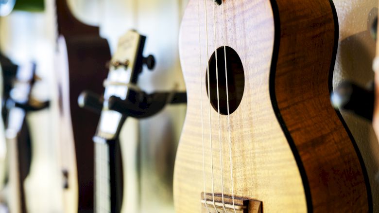 The image shows a close-up of a ukulele hanging on a wall, with other string instruments blurred in the background.