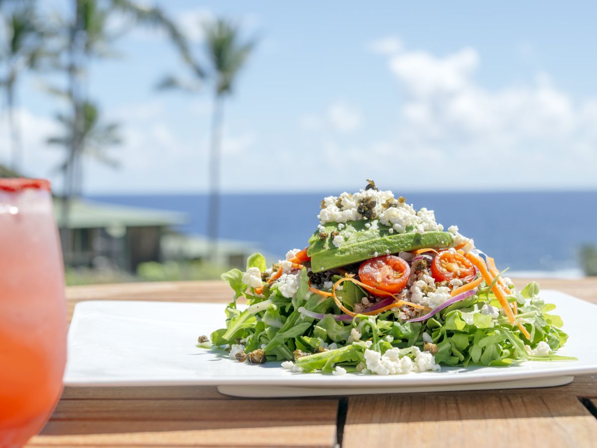 A fresh salad with greens, sliced vegetables, and cheese on a plate, set outdoors with an ocean view and a drink on the side.