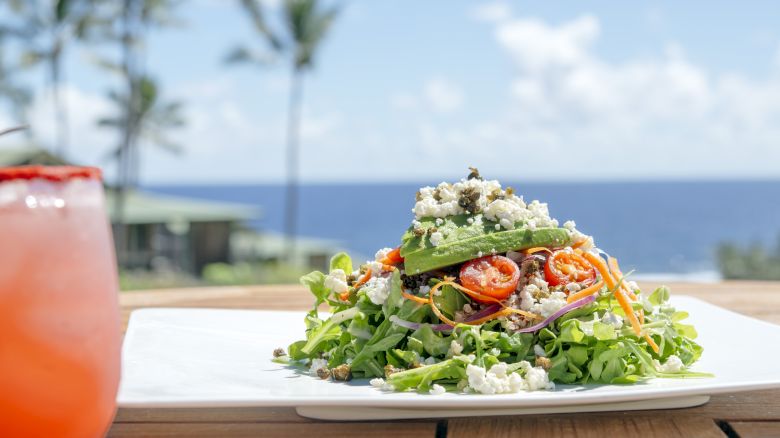 A fresh salad with greens, sliced vegetables, and cheese on a plate, set outdoors with an ocean view and a drink on the side.