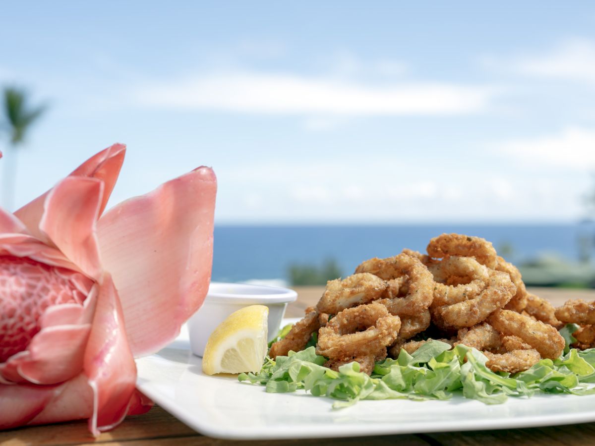 A plate of fried calamari with lemon and sauce, set on a table with a tropical flower, overlooking the ocean and blue sky.