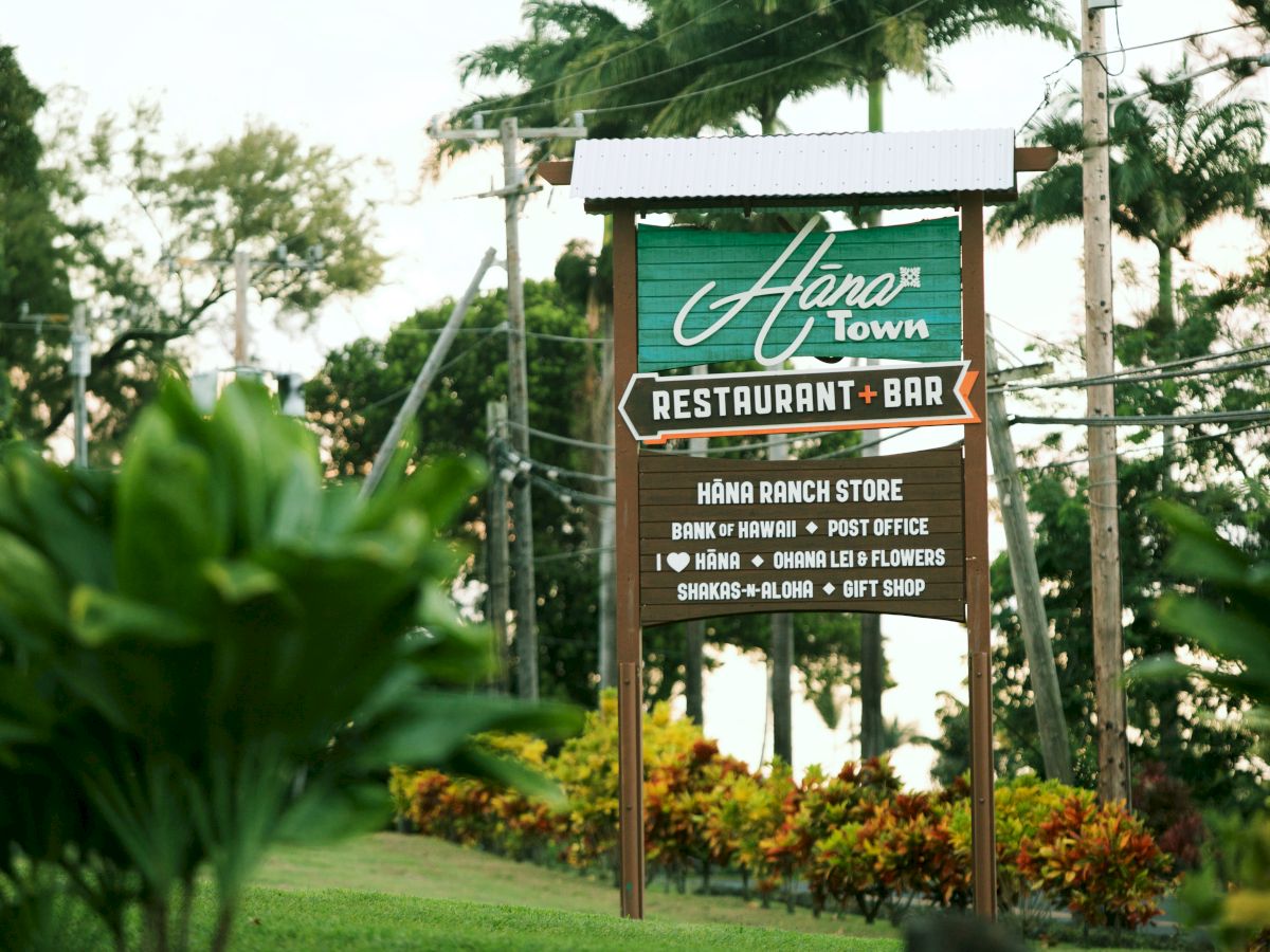 Sign for "Hāna Town Restaurant and Bar" with details about the Hāna Ranch Store, located in a lush, tropical area with greenery and palm trees.