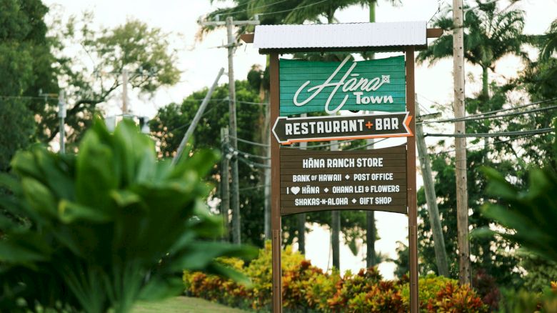 Sign for "Hāna Town Restaurant and Bar" with details about the Hāna Ranch Store, located in a lush, tropical area with greenery and palm trees.