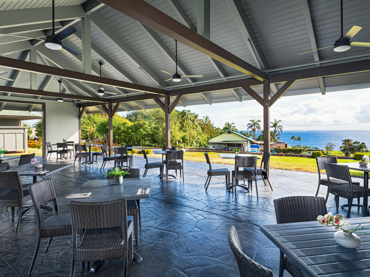 An open-air dining area with wicker furniture, overlooking a scenic landscape with greenery and ocean views in the background.
