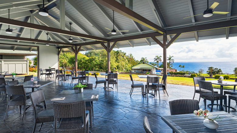 An open-air dining area with wicker furniture, overlooking a scenic landscape with greenery and ocean views in the background.