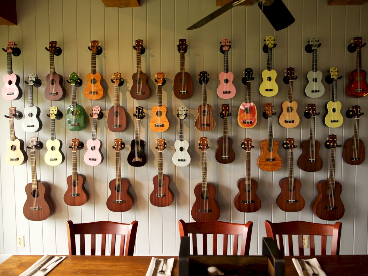 The image shows a wall with a neat, colorful display of numerous ukuleles, and a wooden dining table with chairs and place settings below them.