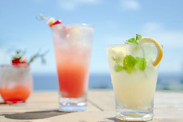 The image shows three refreshing cocktails with garnishes, set against a bright blue sky and water backdrop.