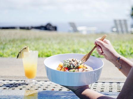 A person holding a bowl of food with a fork, outdoors near a pool. A tropical drink with a pineapple garnish sits nearby on the ledge.