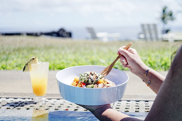 A person holding a bowl of food with a fork, outdoors near a pool. A tropical drink with a pineapple garnish sits nearby on the ledge.