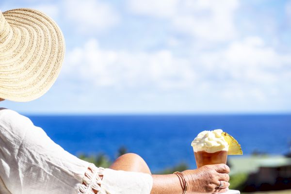 A person in a sun hat enjoys a tropical drink with whipped cream and a pineapple slice, overlooking a scenic ocean view.