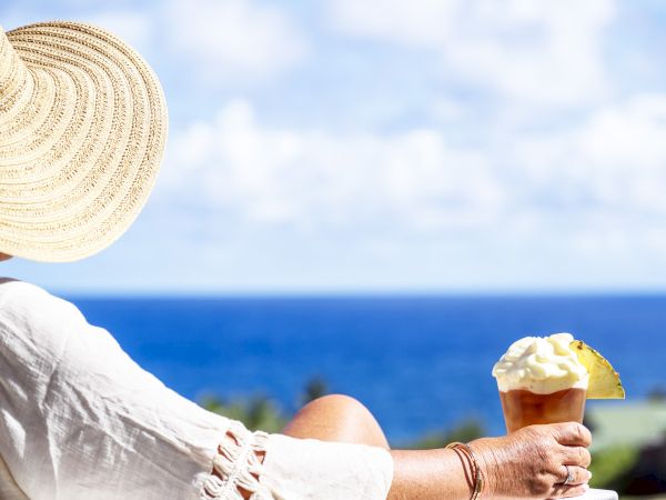 A person in a sun hat enjoys a tropical drink with whipped cream and a pineapple slice, overlooking a scenic ocean view.