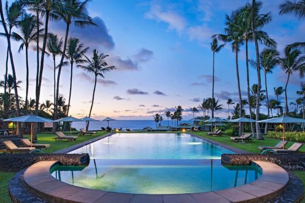 A serene pool surrounded by palm trees with lounge chairs and umbrellas, against a backdrop of a stunning sunset sky over the ocean.