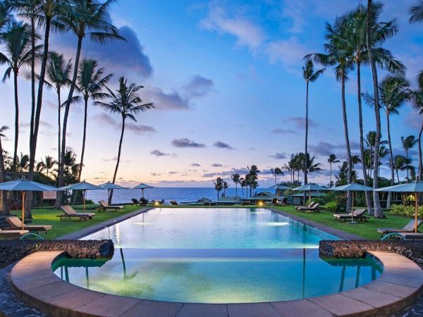 A serene pool surrounded by palm trees with lounge chairs and umbrellas, against a backdrop of a stunning sunset sky over the ocean.