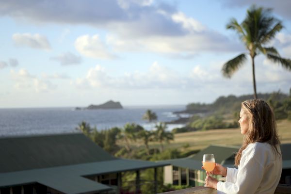 A person with a drink enjoys a scenic ocean view at a tropical location with palm trees and distant island visible under a partly cloudy sky.