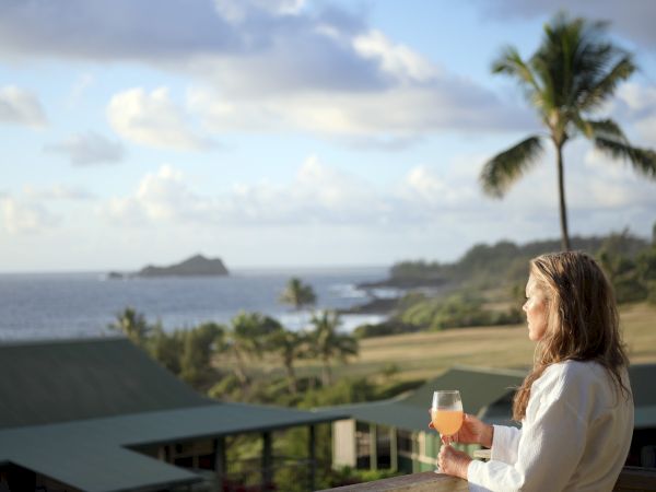 A person with a drink enjoys a scenic ocean view at a tropical location with palm trees and distant island visible under a partly cloudy sky.