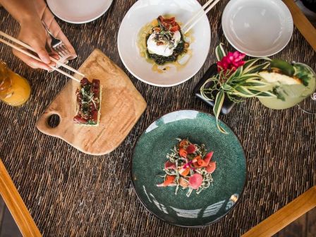 The image shows a table with three beautifully plated dishes, a floral drink, and some hands using chopsticks to pick food, exuding a fine dining setting.