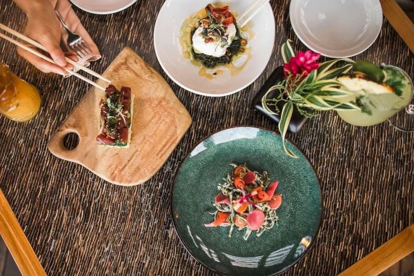 The image shows a table with three beautifully plated dishes, a floral drink, and some hands using chopsticks to pick food, exuding a fine dining setting.