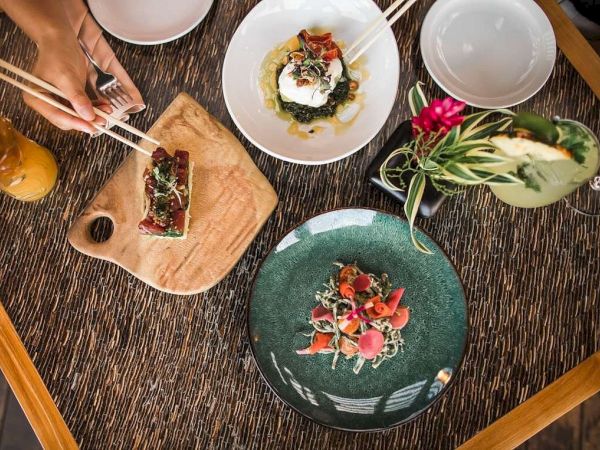 The image shows a table with three beautifully plated dishes, a floral drink, and some hands using chopsticks to pick food, exuding a fine dining setting.