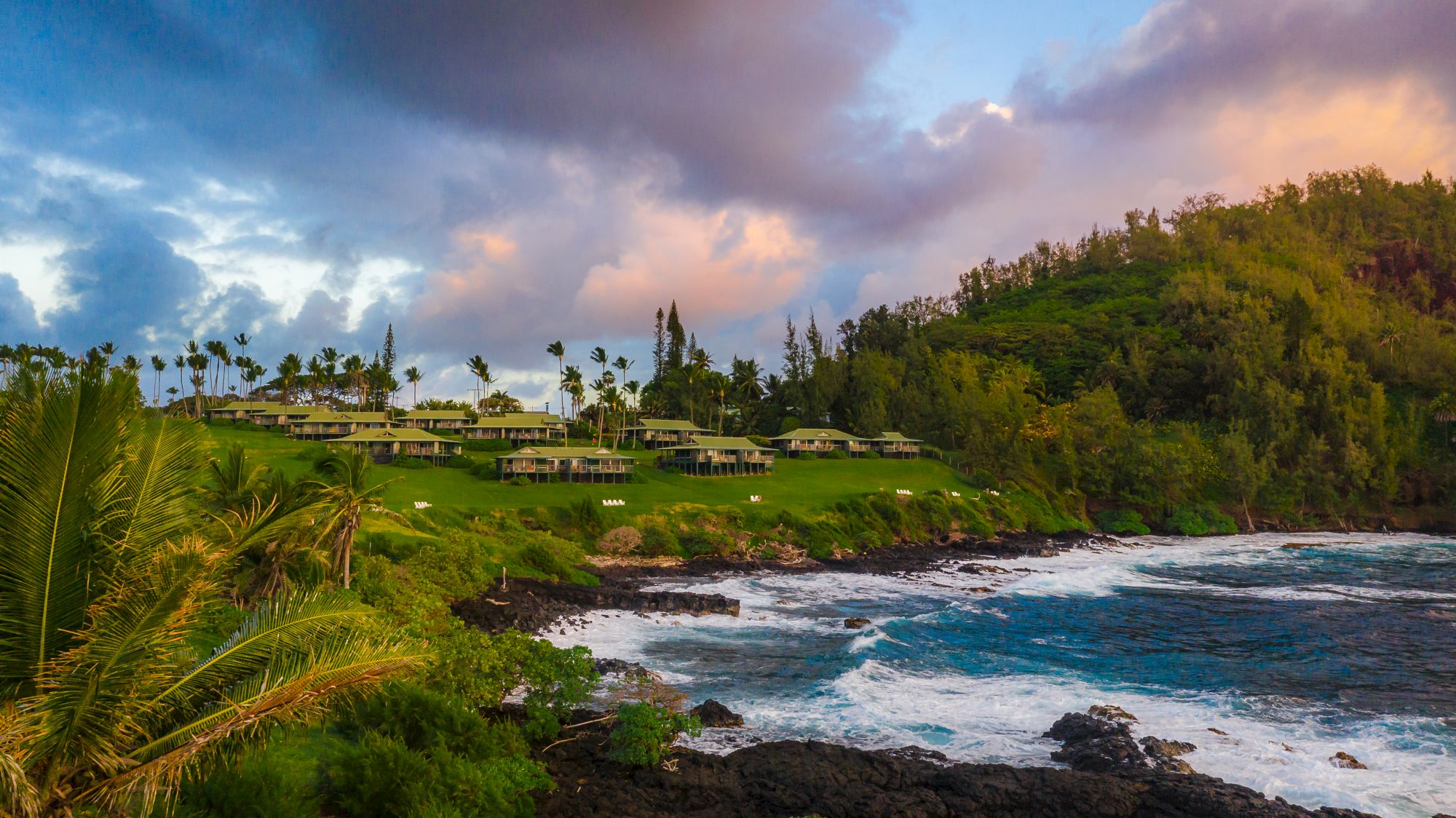 The image shows a coastal resort surrounded by lush greenery with cottages, palm trees, and dynamic ocean waves under a colorful sky at sunset.