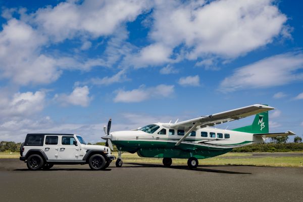 A white SUV is towing a small green and white airplane on a tarmac under a partly cloudy sky.