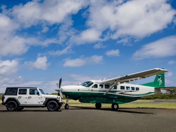 A white SUV is towing a small green and white airplane on a tarmac under a partly cloudy sky.
