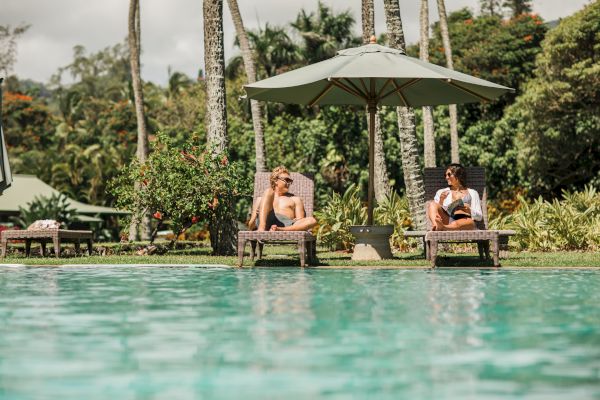 Two individuals relax on lounge chairs by a pool, shaded by an umbrella, surrounded by lush greenery and tall palm trees.