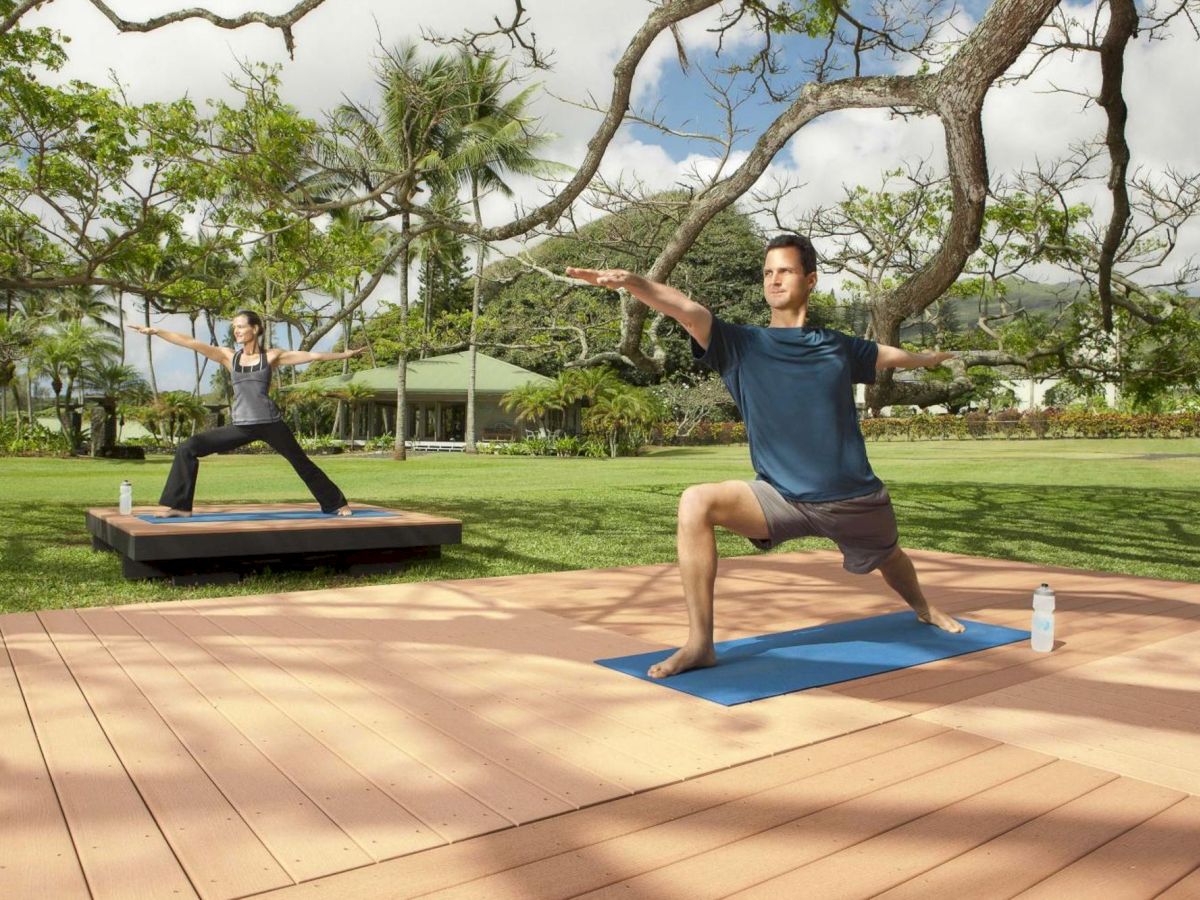 Two people are practicing yoga outdoors on mats in a scenic garden with trees and mountains in the background, with water bottles nearby.